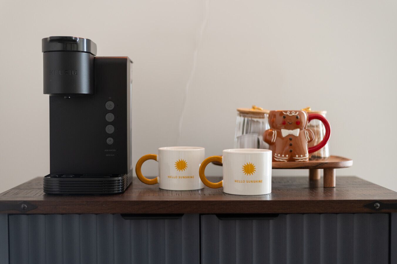 Una cafetera, dues tasses amb la inscripció “Hello Sunshine” i una tassa amb forma de noi de melmelada sobre una taula de fusta.