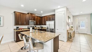 A modern kitchen with dark wooden cabinets, a stainless steel refrigerator, a granite countertop, and a built-in island with bar stools. - New 8BR Luxury Retreat • Pool & Spa • Near Disney (Kissimmee)