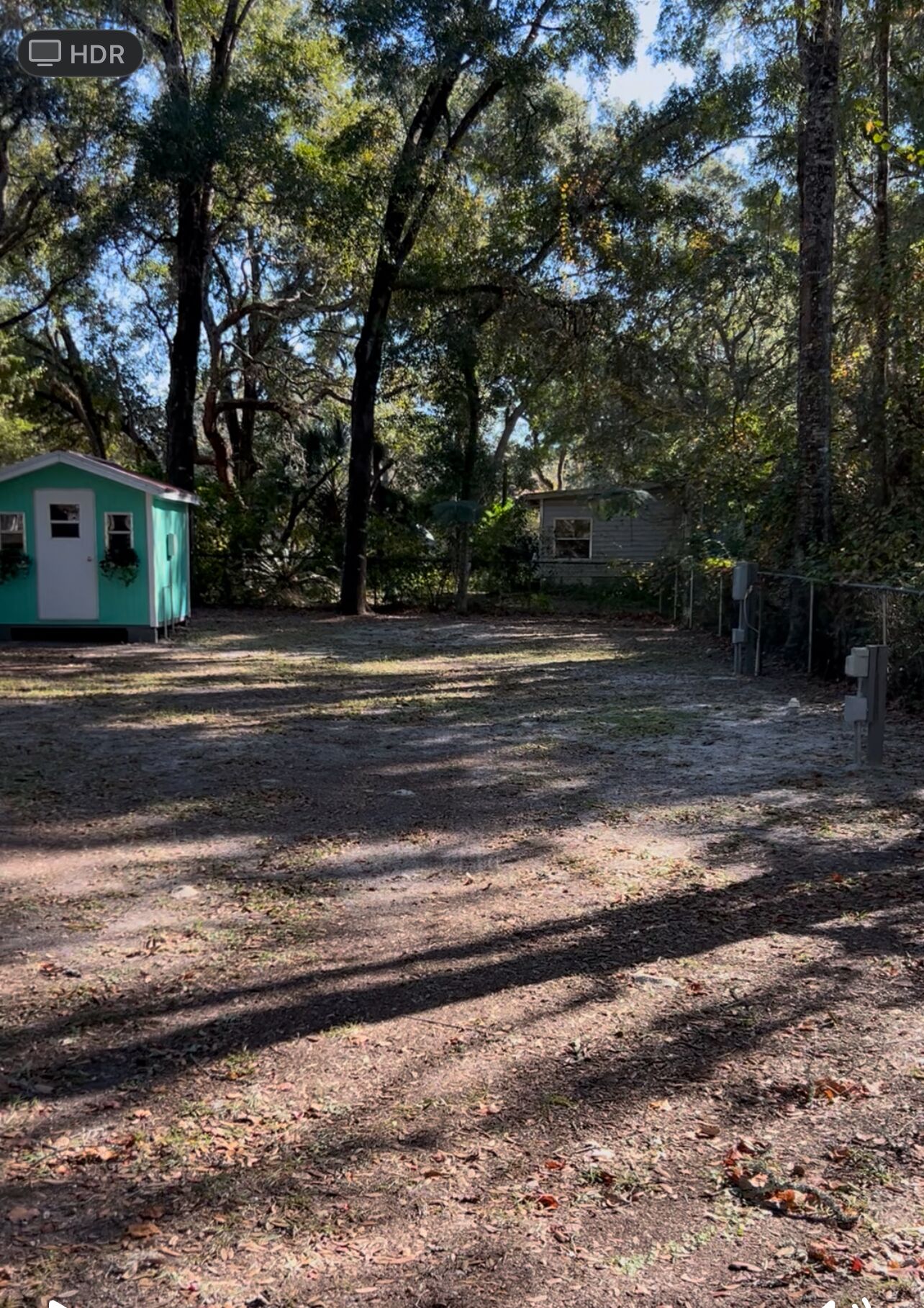 Une zone boisée avec un petit bâtiment vert, qui pourrait être une cabane ou un abri de jardin, et une zone délimitée par une clôture, où se trouvent des boîtiers techniques.