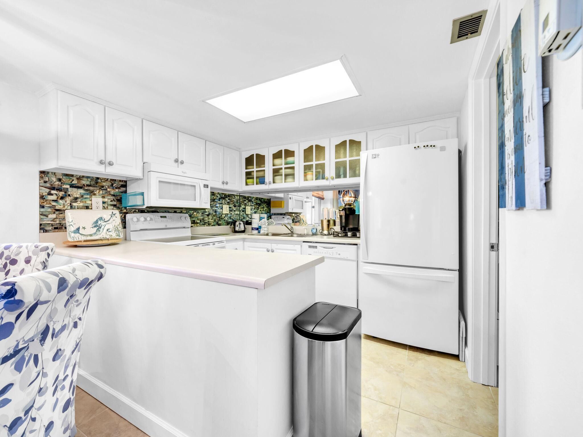 A modern kitchen with white cabinets, a central island, a microwave, a refrigerator, and a trash bin.
