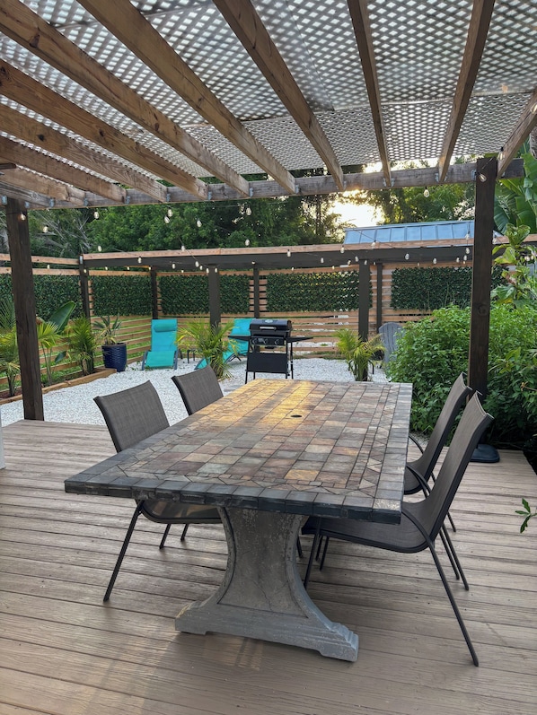 An outdoor patio with a wooden table and chairs, a pergola with a metal roof, and a green hedge in the background.