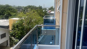 A balcony with a glass railing, a tiled floor, and a view of the surrounding area including trees and buildings.