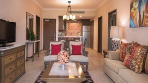 A living room with a TV, sofa, and coffee table. There is a kitchen in the background with a refrigerator and cabinets. - 2-bedroom resort in fantastic Loreto with AC and fitness room (Loreto)