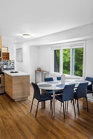 Una cocina moderna con una isla central, un comedor con una mesa redonda y sillas, y un amplio ventanal con vistas al verdor.