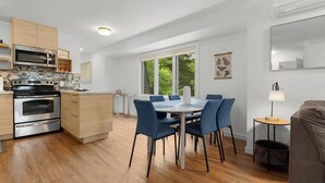 Una cocina moderna con electrodomésticos integrados, un comedor con sillas azules y amplios ventanales con vistas al verdor.