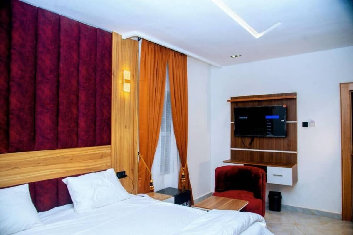 A hotel room with a bed, a TV mounted on the wall, a red chair, and a wooden headboard.