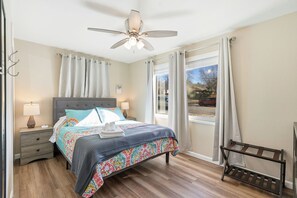 Un dormitorio con cama, mesitas de noche, ventilador de techo, ventana con cortinas y vistas al exterior.