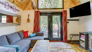 A cozy living room with a grey sofa, red and blue cushions, a mounted TV, and a large window with curtains.