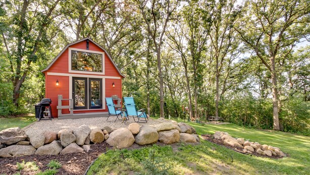 A red cabin with a large window, two blue chairs, and a stone wall in a wooded area.