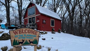 A wooden sign welcoming visitors to a playhouse in the woods, with a red building in the background.