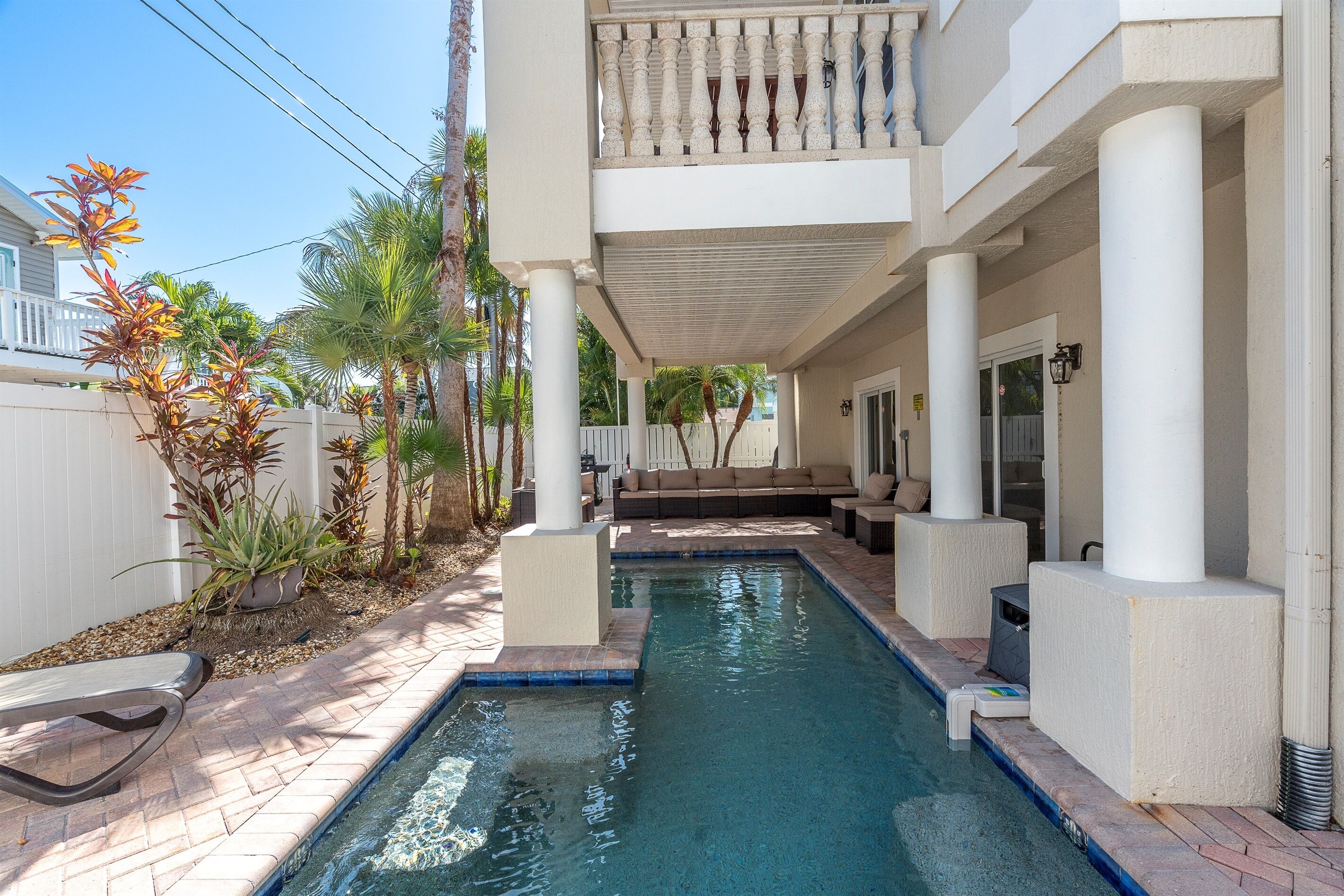 A swimming pool with a covered patio area, a white fence, and palm trees.