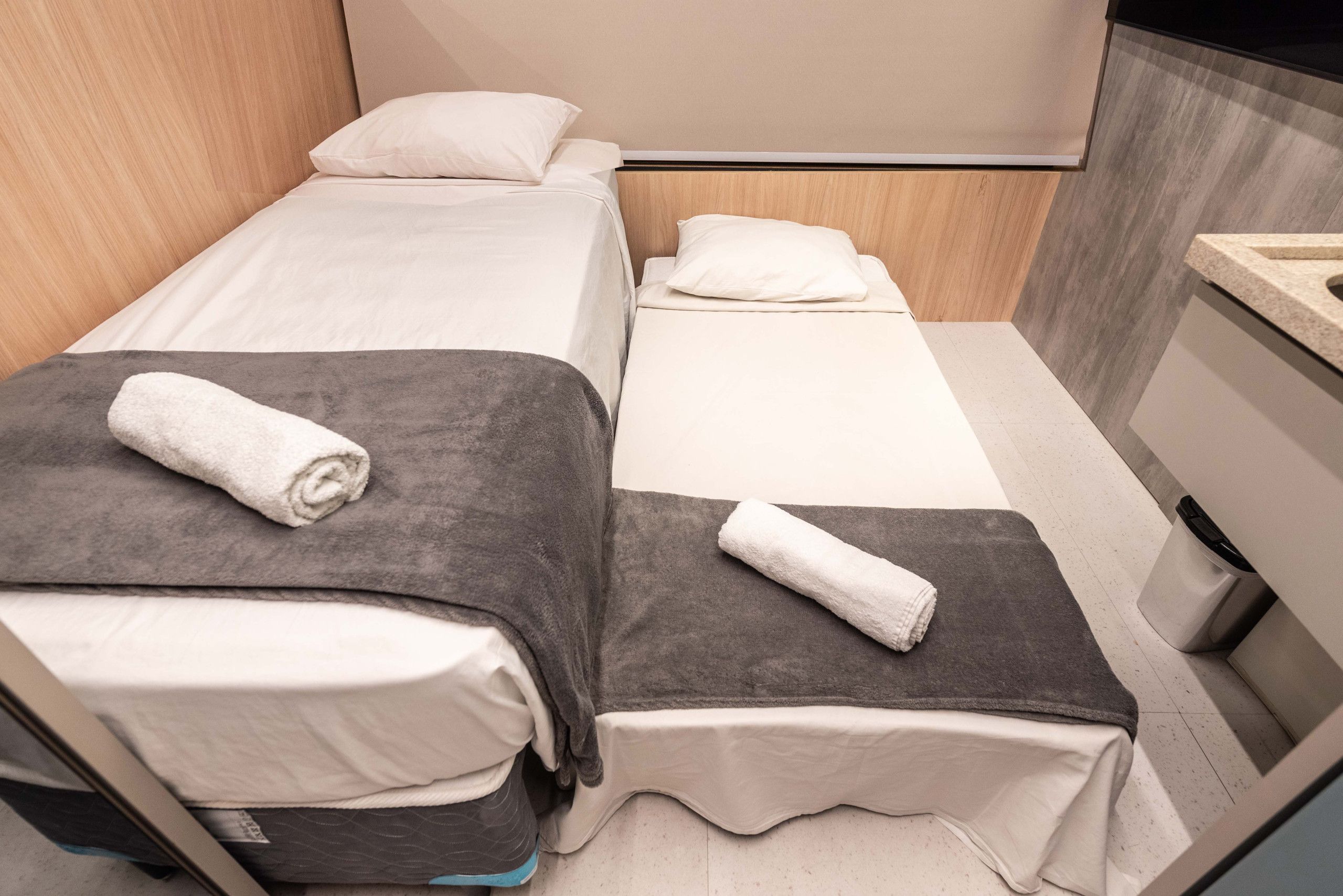 A hotel room with two single beds, each with a white mattress and a grey blanket, and a small bedside table with a lamp.