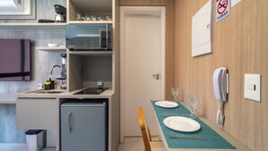 A compact kitchen with a microwave, coffee maker, and a small refrigerator. A dining table with a placemat and a wine glass is set up. - Melo Alves_31 2p / 1q (São Paulo)