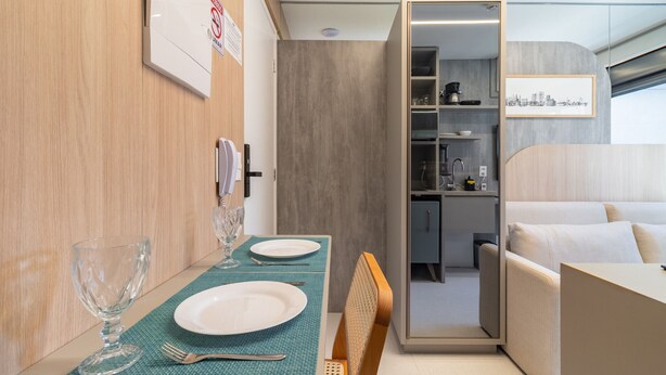 A compact, modern interior with a dining area featuring a table set with plates, cutlery, and a glass. A sofa and a small kitchenette are visible in the background. - Melo Alves_31 2p / 1q (São Paulo)