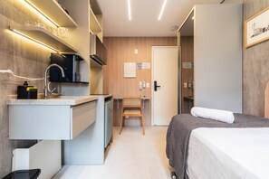 A compact hotel room with a kitchenette, a bed, a chair, and a door with a sign. - Melo Alves_31 2p / 1q (São Paulo)