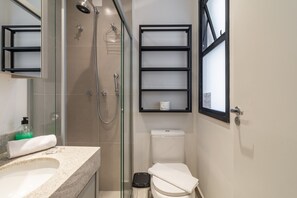 Ein modernes Badezimmer mit Duschkabine aus Glas, einer weißen Toilette, einem Waschbecken mit Ablagefläche und einem schwarzen Mülleimer.