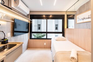 Ein kompaktes Hotelzimmer mit einem Bett, einer kleinen Küchenzeile, einem an der Wand befestigten Fernseher und einem Fenster mit Blick auf Gebäude.