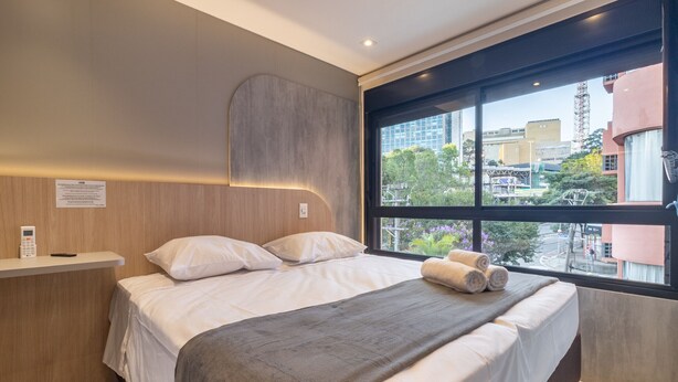 Ein modernes Hotelzimmer mit einem großen Bett, einem Nachttisch mit Telefon und einem Fenster mit Blick auf die Stadt.