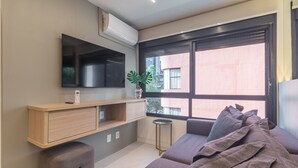 Ein modernes Wohnzimmer mit Flachbildfernseher, einem Holzkonsole, einem kleinen Hocker, einer Couch mit Kissen und einem großen Fenster mit Blick auf Gebäude.