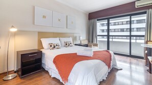 Hotellihuone, jossa on suuri sänky, yöpöytä, lattiavalaisin ja suuri ikkuna, josta avautuu näkymä rakennukseen.