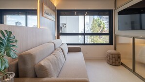 Un salon moderne avec un canapé beige, un petit pouf rond, une grande fenêtre donnant sur la ville et une plante placée à gauche.