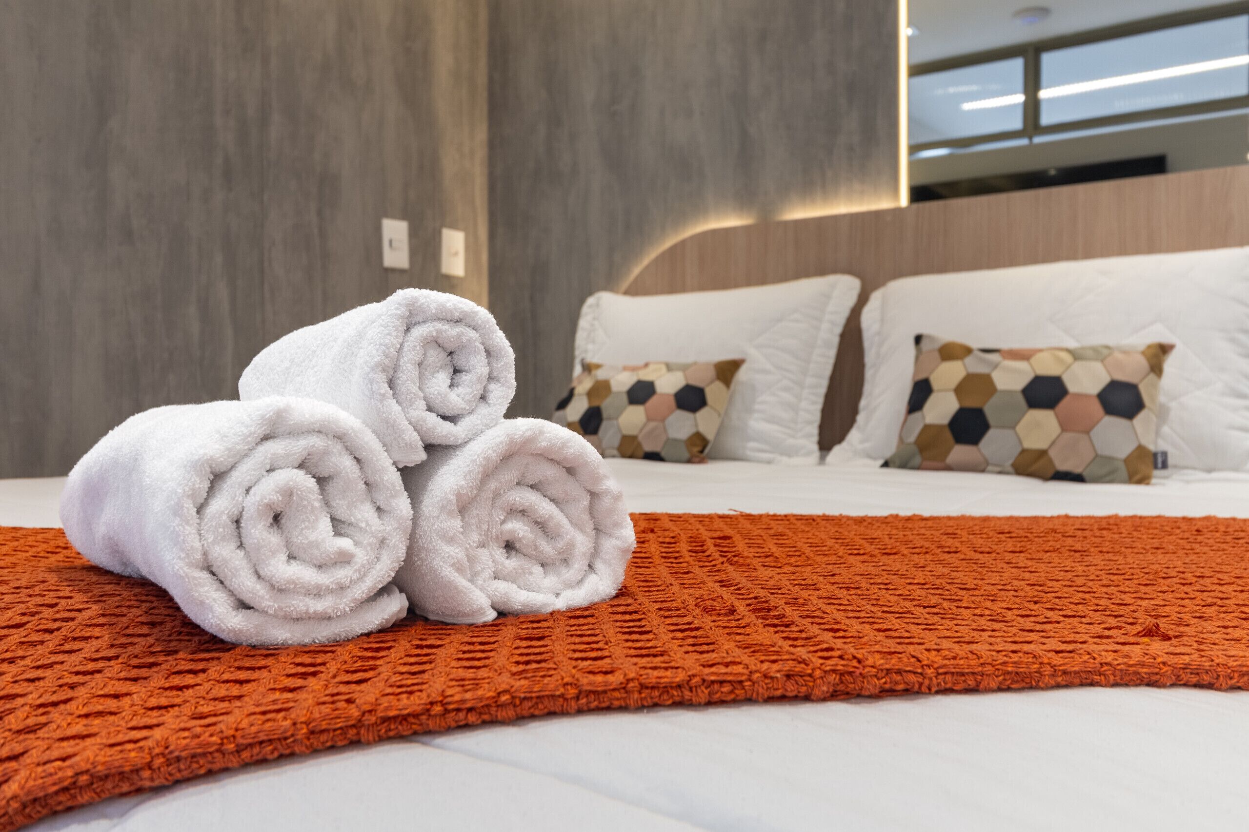 Una cama bien tendida con toallas blancas enrolladas y una manta naranja texturizada, almohadas blancas con un patrón hexagonal y un cabecero de madera.