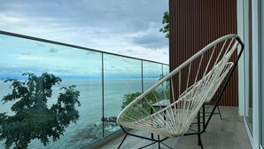 A balcony with a white wicker chair, a glass railing, and a view of a body of water and distant land. - Luxury condo with Rooftop pool & Ocean views (Boca de Tomatlán)