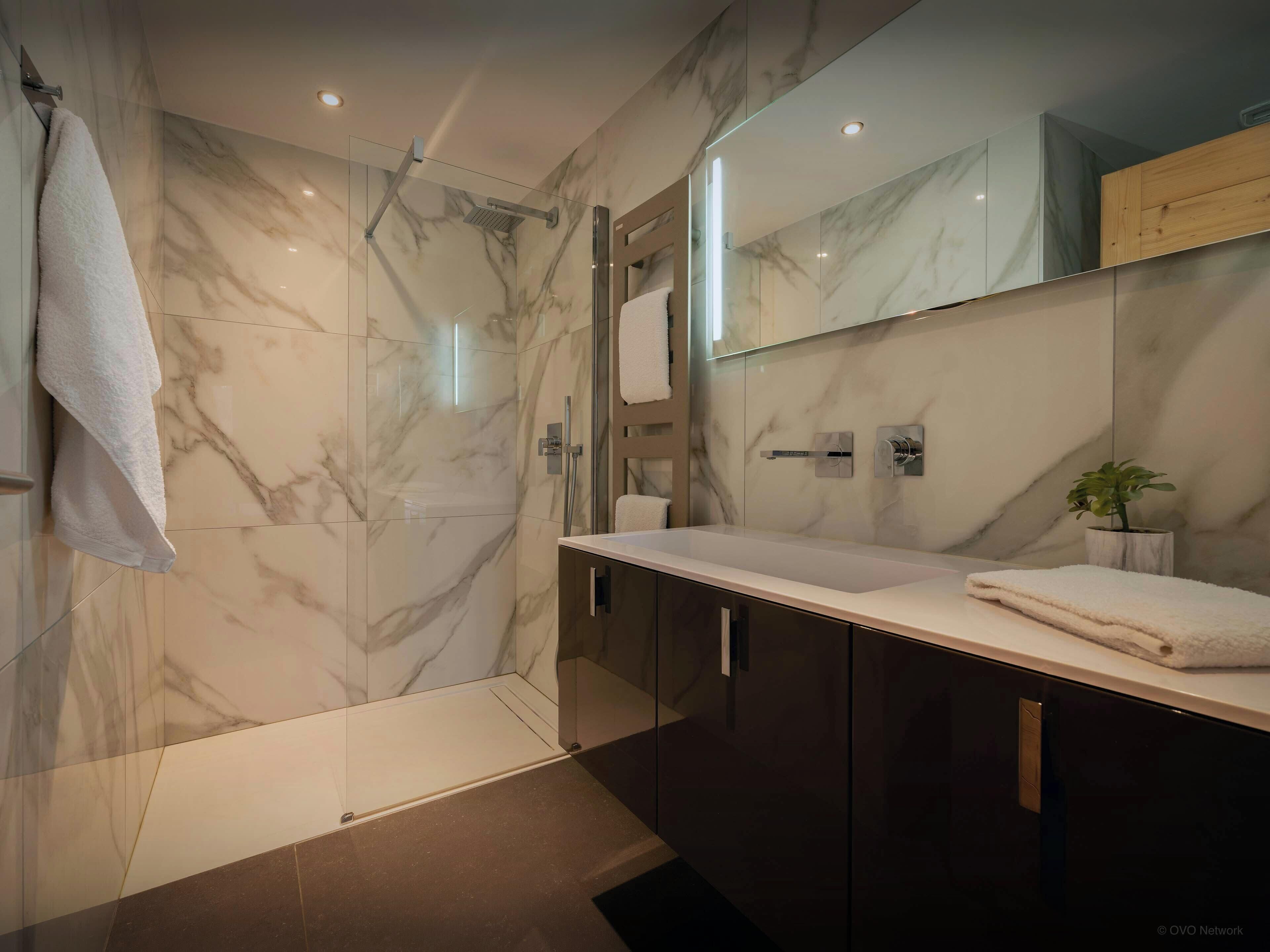 A modern bathroom with a glass-enclosed shower, marble walls, a large mirror, and dark wooden cabinets.