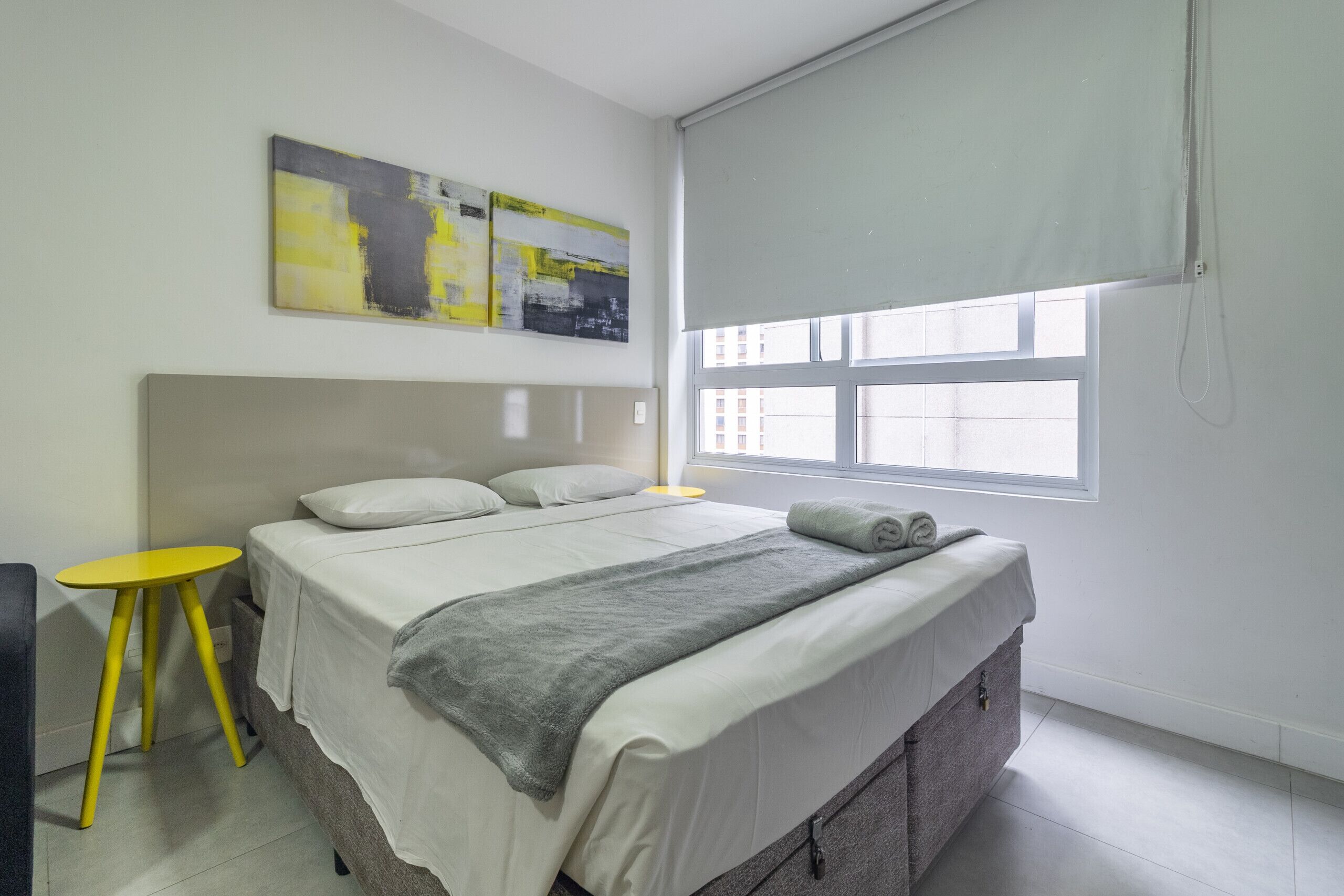 Un dormitorio con una cama, una mesita amarilla pequeña y una manta gris, y una ventana con persianas.