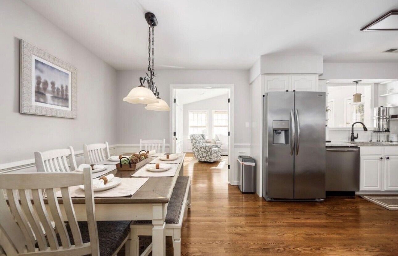 A modern kitchen with white cabinetry, a stainless steel refrigerator, and a dining area with a wooden table and white chairs.
