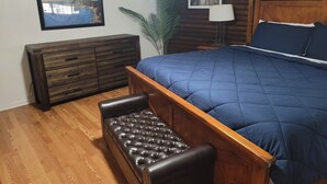 Ein Schlafzimmer mit hölzernem Kopfteil, einer blauen gesteppten Bettdecke, einer dunklen Lederbank, einem Holzkleiderschrank und einem gerahmten Bild an der Wand.