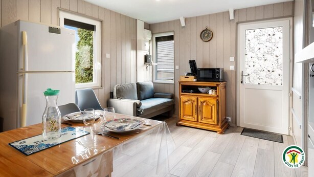 A compact kitchen with a refrigerator, microwave, and wooden cabinets. There is a dining table set with plates and glasses. - Gîtes de France® - La Maison au Calme (CRIEL SUR MER)