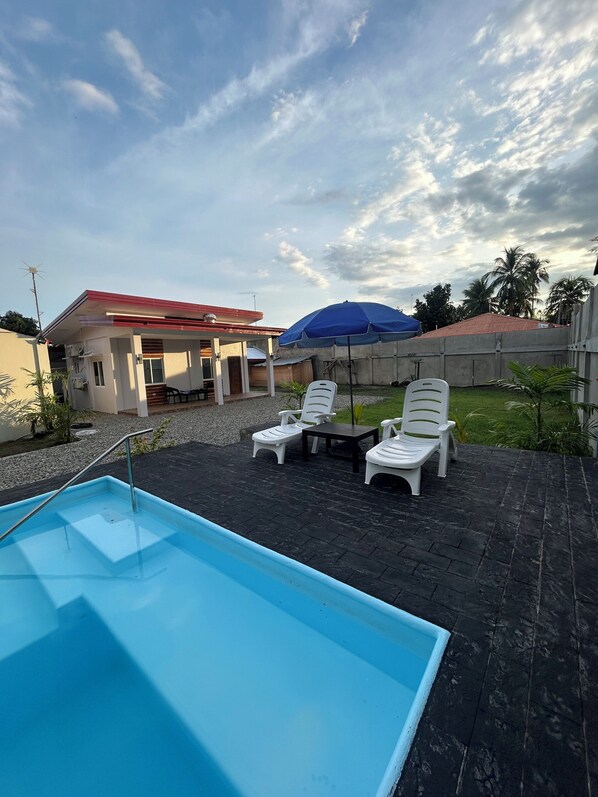 A swimming pool with lounge chairs and a covered area, surrounded by a garden and a building with a red roof.