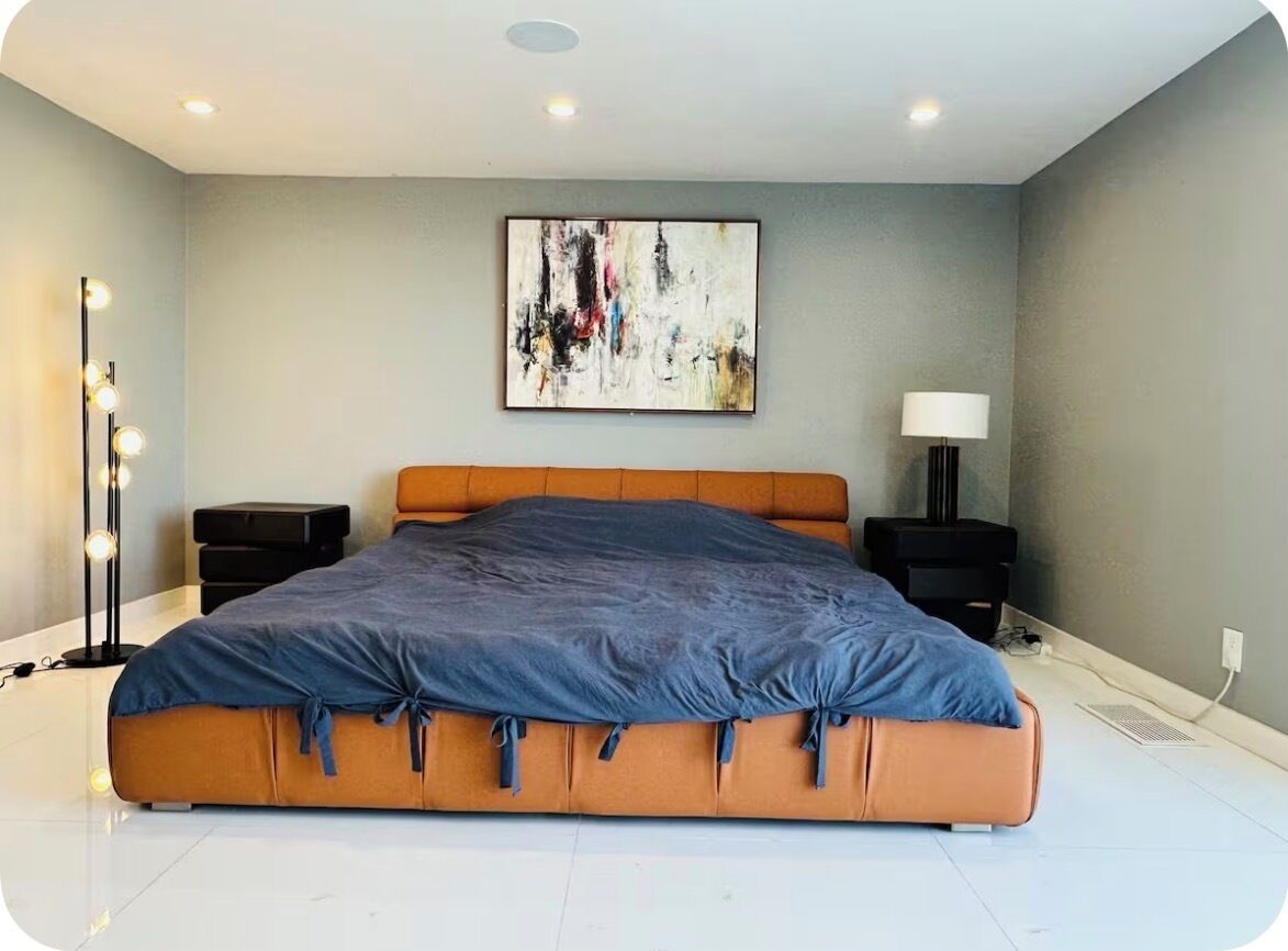 A modern bedroom with a large bed, a floor lamp, a side table, and a wall-mounted abstract painting.