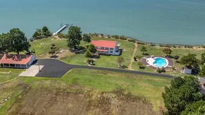 Vista aérea de uma propriedade costeira com uma casa grande, piscina e um cais que se estende para a água.