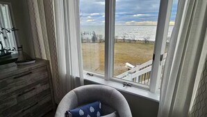 Quarto com uma janela grande, uma poltrona cinza com almofada azul, cômoda de madeira e tapete estampado.