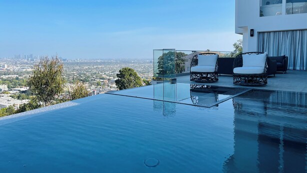 Una piscina infinita en la azotea con vistas a la ciudad, dos sillones blancos acolchados y un área de estar exterior moderna.