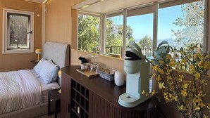 Habitación de hotel con cama, escritorio con cafetera, ventana con vistas a árboles y edificios, y mesita de noche con lámpara.