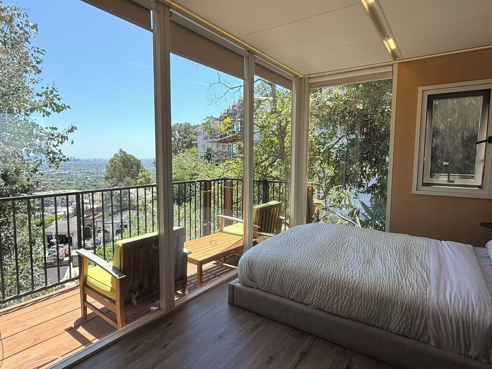 A bedroom with a large bed, a balcony with a wooden table and chairs, and a city view.
