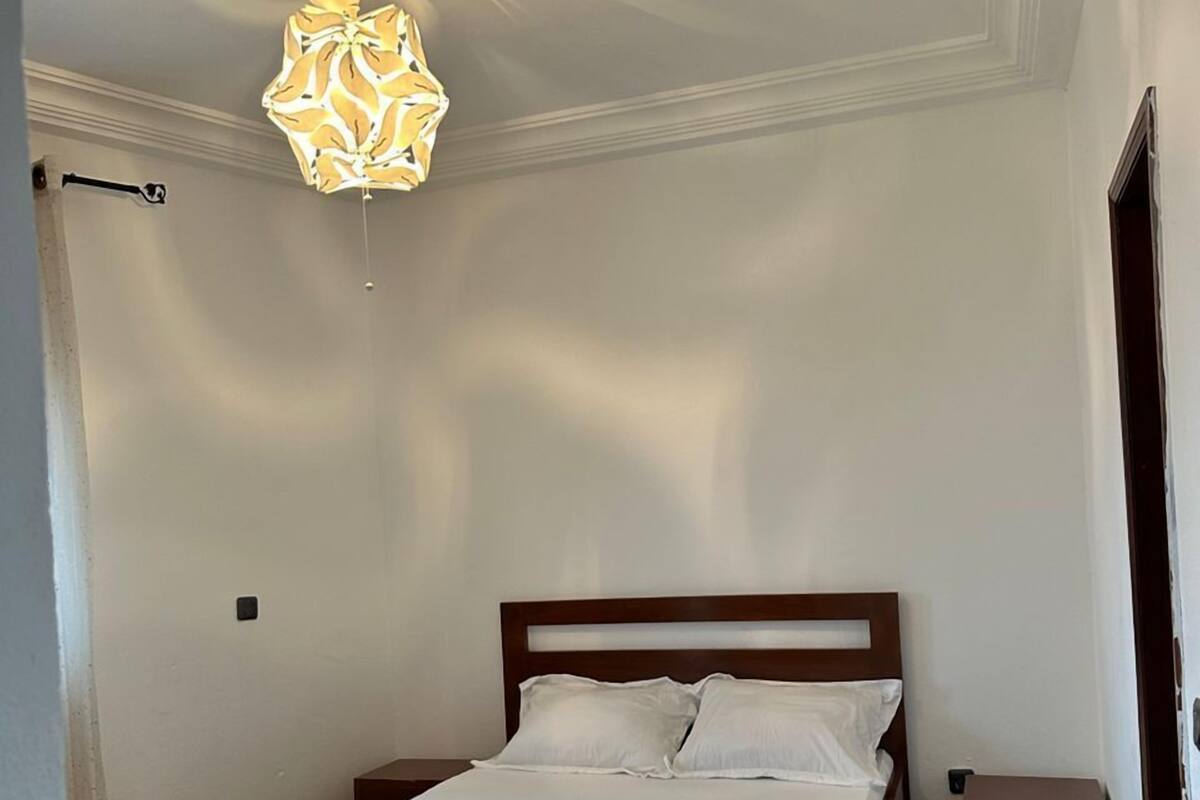A bedroom with a wooden bed, white bedding, a nightstand, and a ceiling light fixture.
