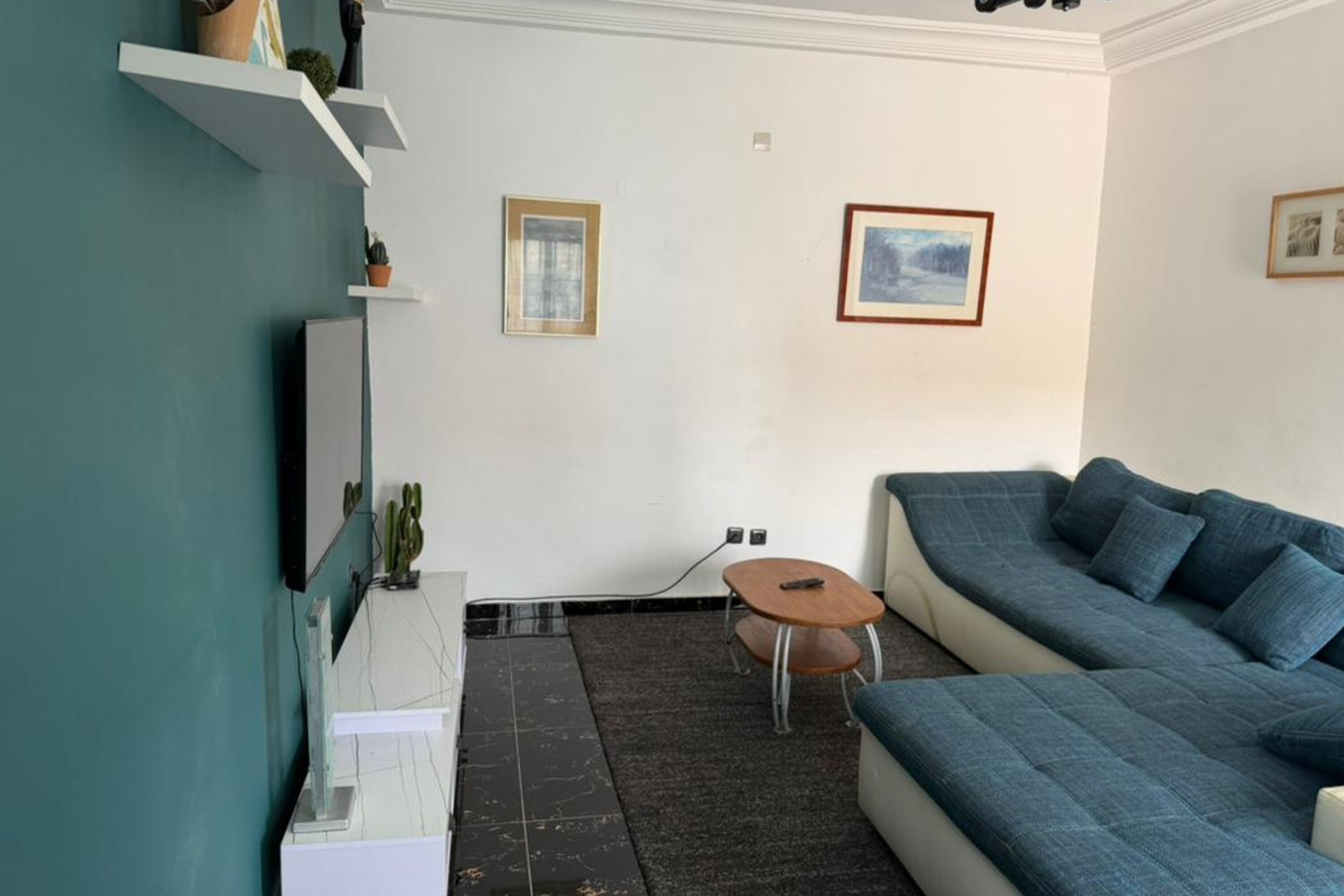 A living room with a blue sofa, a small round table, and a white cabinet. There are two framed pictures on the wall.