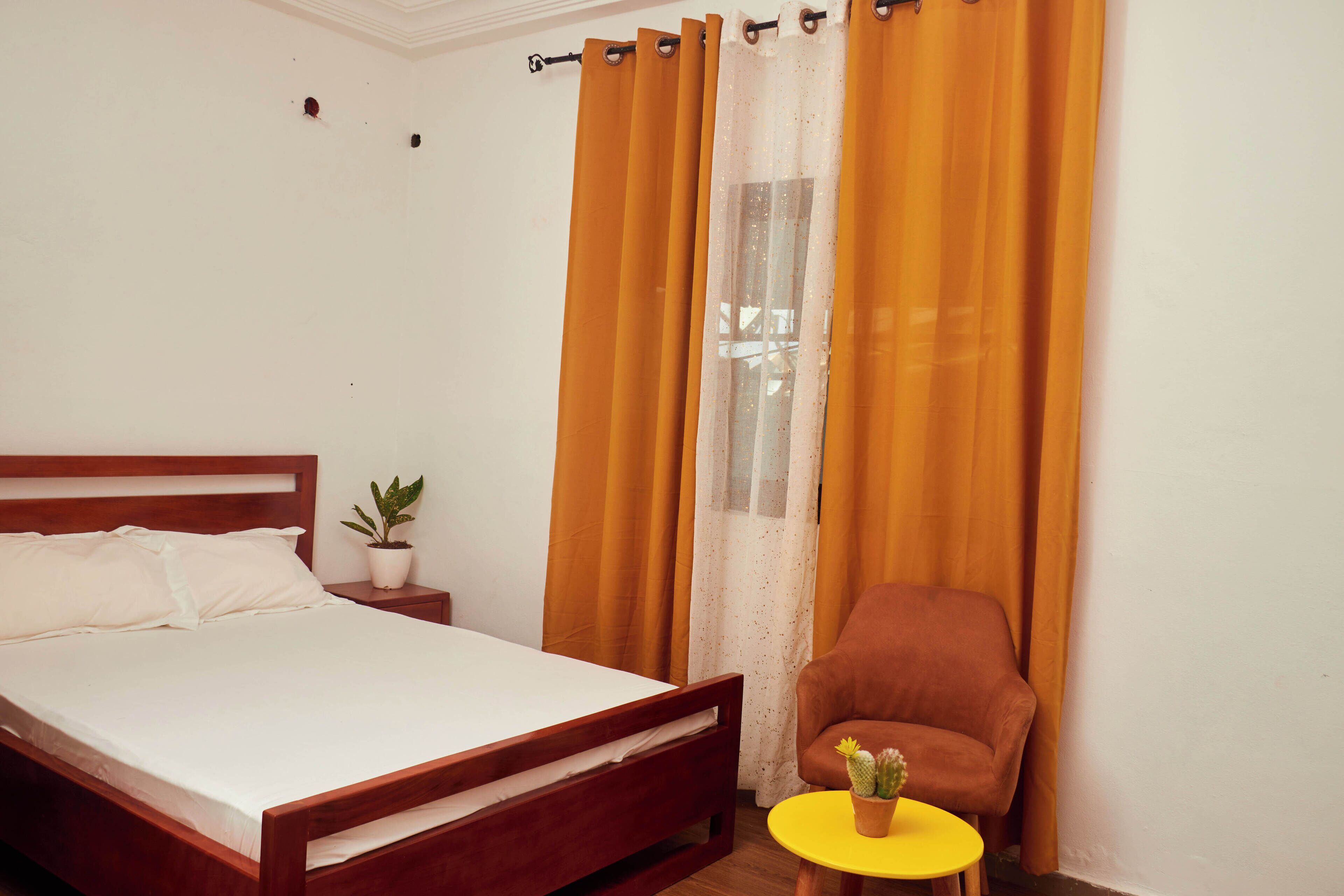 A bedroom with a wooden bed, a small round table with a potted plant, a brown armchair, and orange curtains.