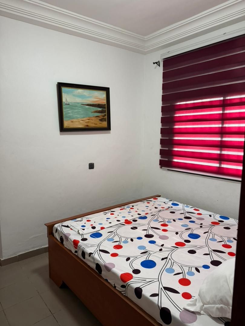 A room with a bed, a painting on the wall, and a window with red blinds.