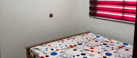 A room with a bed, a painting on the wall, and a window with red blinds.