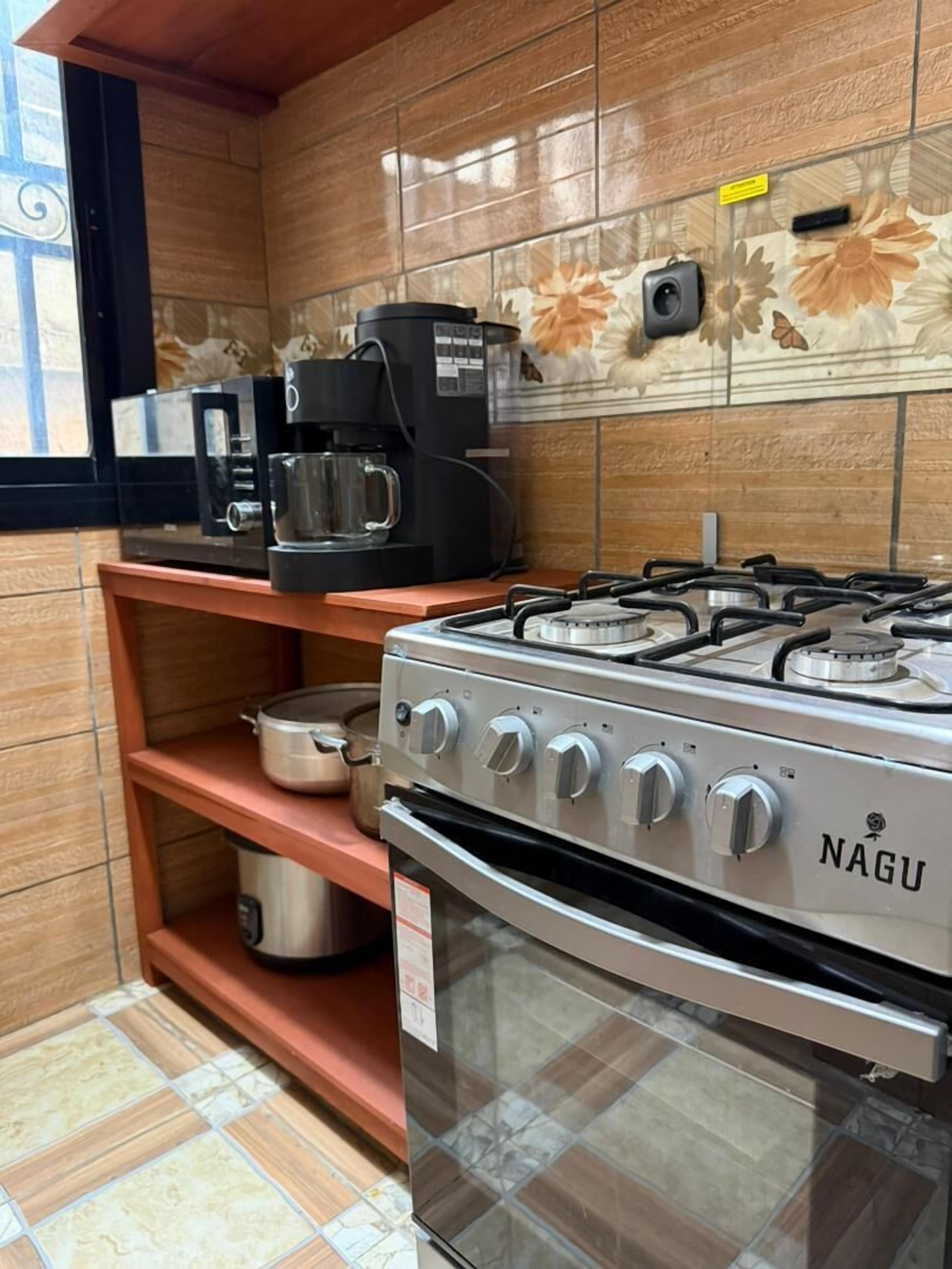 A kitchen with a NAGU gas stove, a coffee maker, and a shelf with pots.