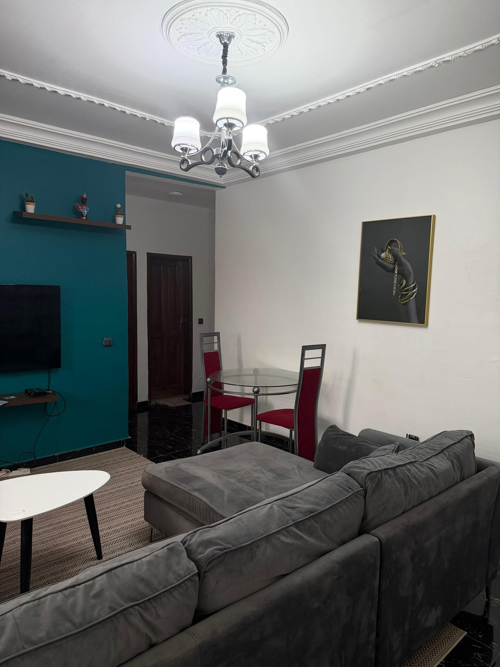 A living room with a grey sofa, a glass table, red chairs, a TV on the wall, and a chandelier.