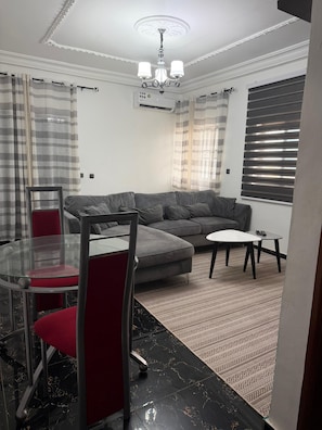 Ein Wohnzimmer mit einem Ecksofa, einem Glastisch mit roten Stühlen, einem gemusterten Teppich, einer Deckenleuchte und einem Fenster mit Jalousien.