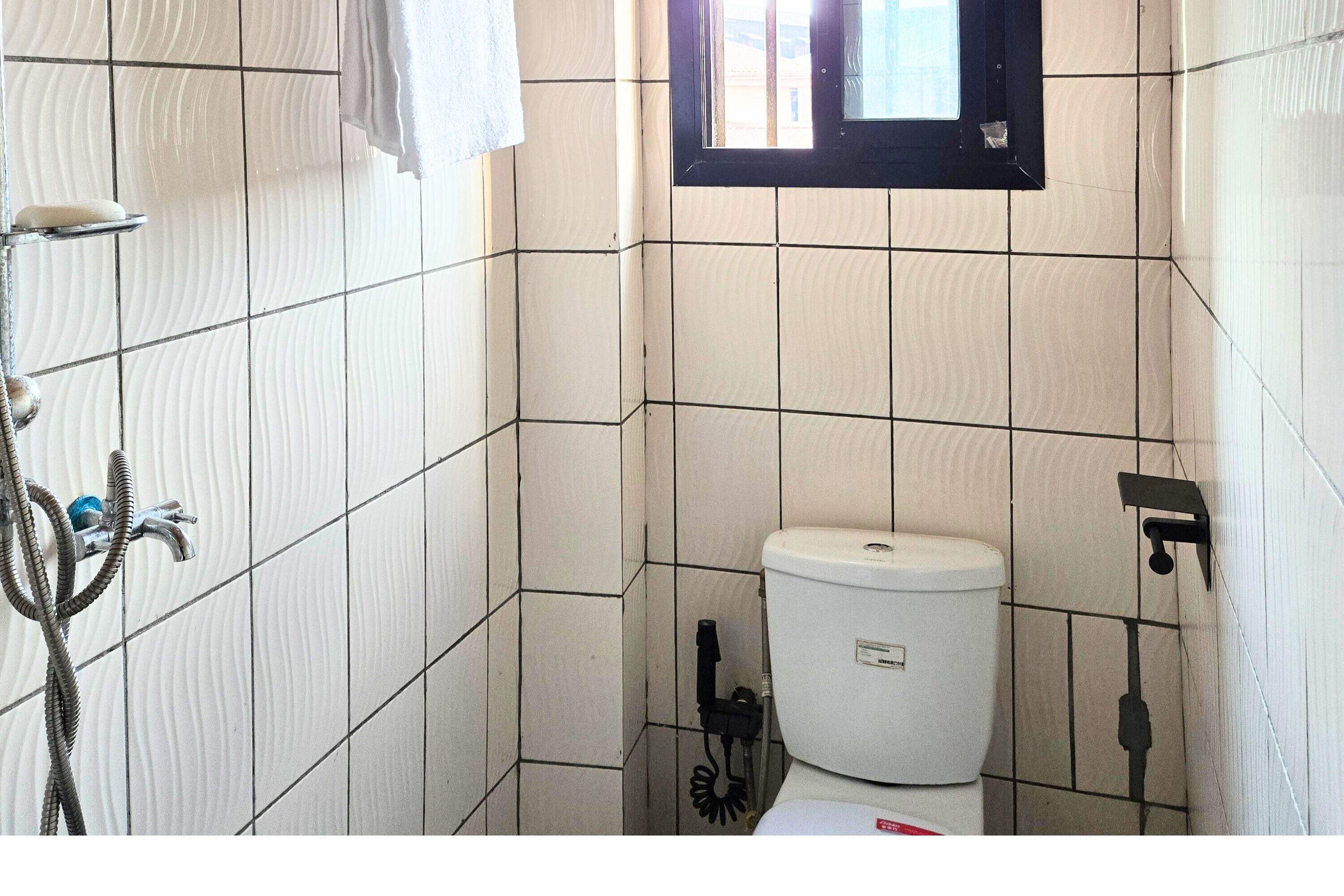 Een kleine badkamer met witte betegelde wanden, een toilet, een douche en een raam.