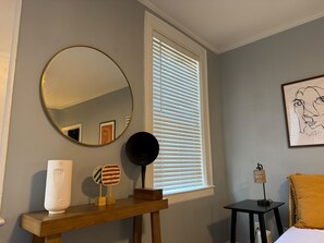 Ein Zimmer mit einem Holzkonsolentisch, einem runden Spiegel, einem gerahmten Kunstwerk, einem Fenster mit Jalousien und einem Bett mit einem gelben Kissen.