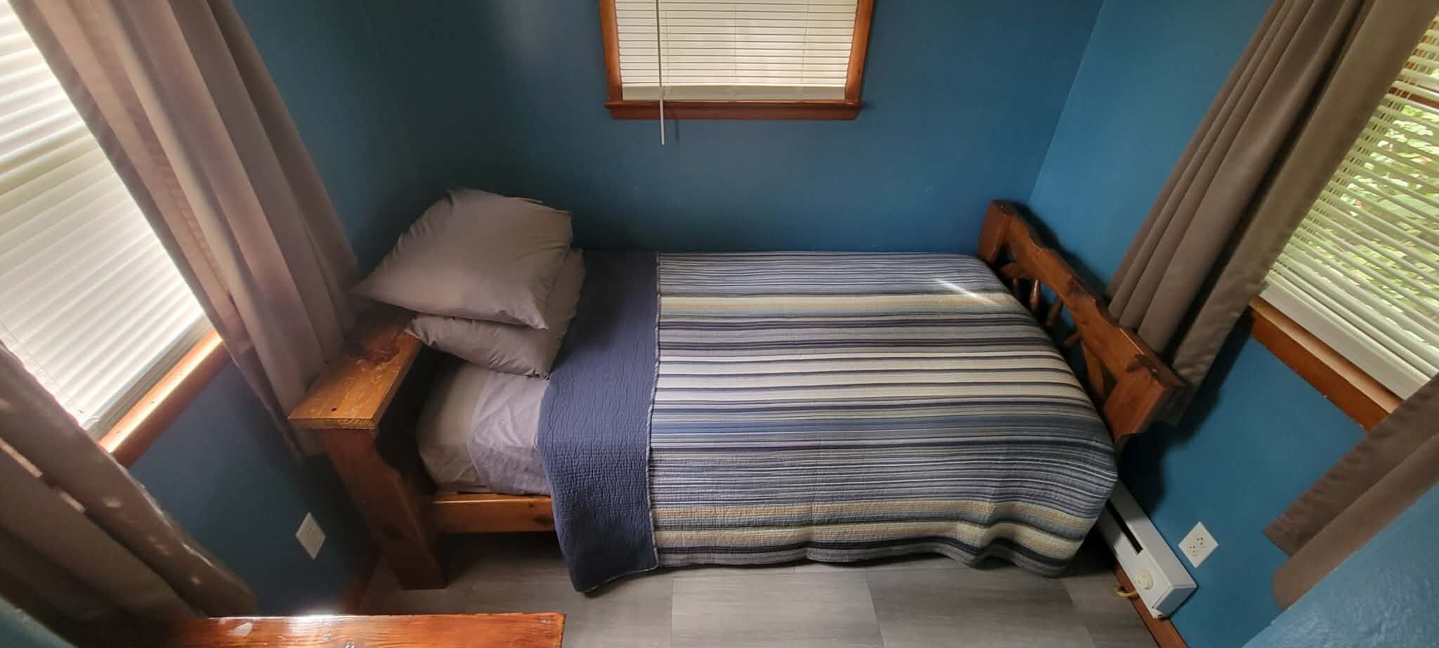 Ein kleines Zimmer mit einem Einzelbett, einem Holz-Kopfteil, einem Fenster mit Jalousien und einer Bettdecke mit blauen Streifen.